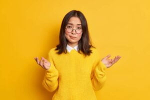 SGP Credit - Personal Loan With Low Interest Rate | Unaware hesitant Asian woman shrugs shoulders in clueless gesture cannot make decision wears round spectacles and casual jumper isolated over yellow background. Indifferent cluess female model
