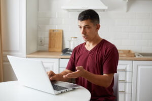 SGP Credit - Personal Loan With Low Interest Rate | Indoor shot of young adult male freelancer sitting in kitchen at white table, working online on computer, having problems with work, looking at pc screen with puzzled expression.