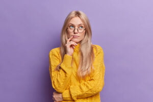 SGP Credit - Personal Loan With Low Interest Rate | Young pretty young woman thinks of ideas concentrated above stands thoughtful and keeps hand on face stands in thoughtful pose wears round glasses yellow sweater isolated over purple background.