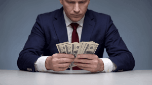 Person in a navy suit counts a stack of US $100 bills on a desk in a neutral setting, focused on money