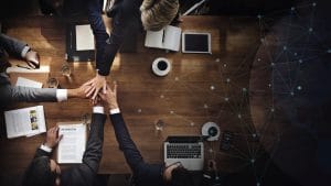 Top-down view of professionals joining hands in the center of a wooden conference table, with notebooks, tablets, and coffee nearby—symbolizing teamwork.