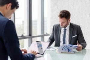 Two business colleagues review documents and a tablet at a glass conference table in a bright office.