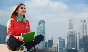 Young woman in a red jacket sits cross-legged on a rooftop with teal headphones, holding a green notebook against a city skyline backdrop.
