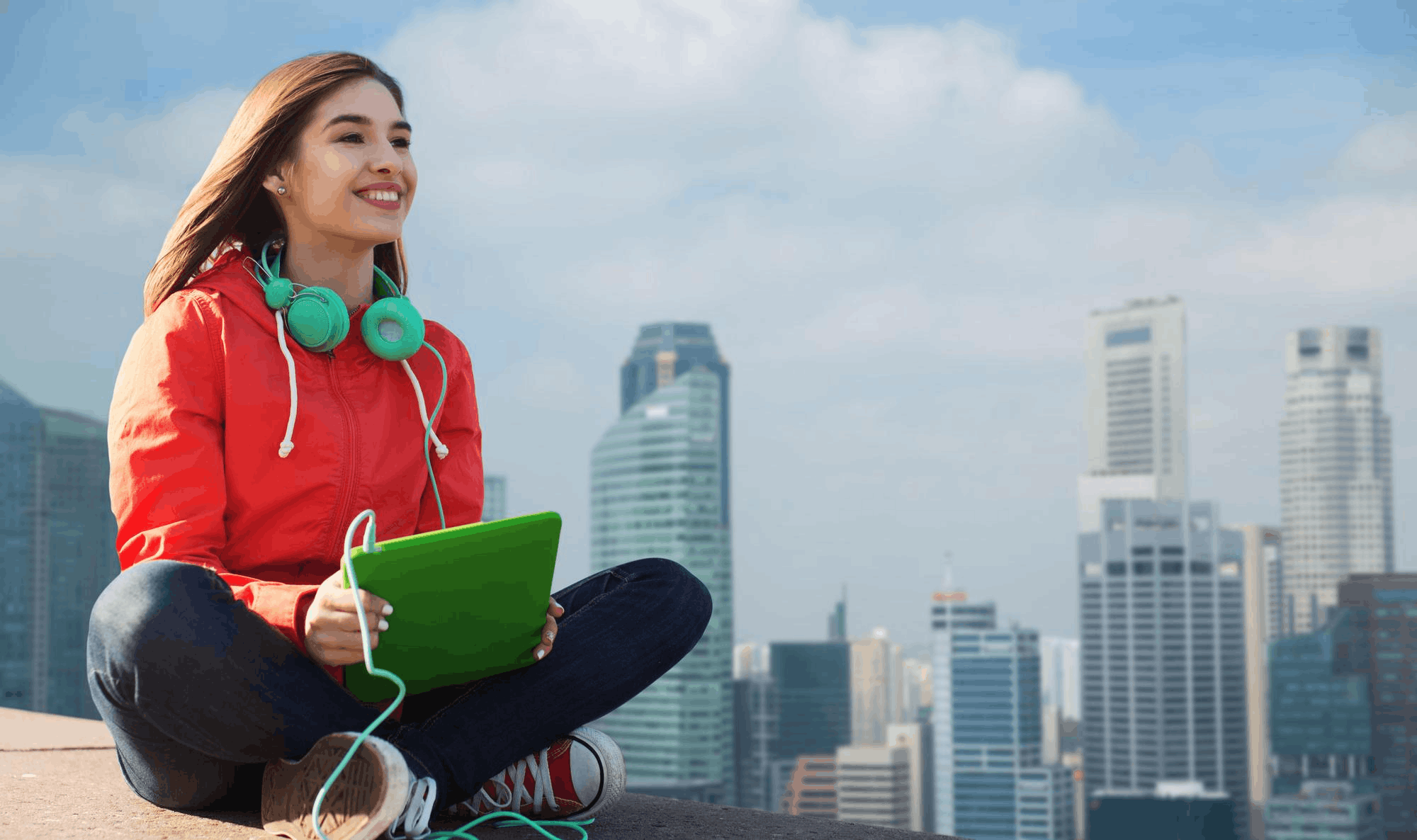 Young woman in a red jacket sits cross-legged on a rooftop with teal headphones, holding a green notebook against a city skyline backdrop.