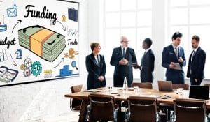 Group of professionals in business attire discussing funding ideas beside a whiteboard filled with money illustrations and charts in a bright conference room.