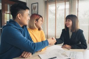 Three coworkers shake hands across a desk in a bright office, with papers and a clipboard visible on the table.