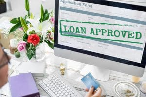 Monitor shows a form on screen with a bold green 'LOAN APPROVED' stamp across it, on a desk with flowers nearby.