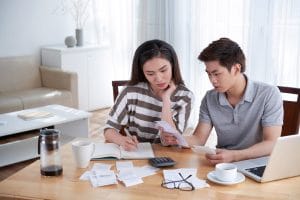 A woman and a man sit at a wooden table surrounded by receipts, papers, and a laptop, looking focused while reviewing documents.