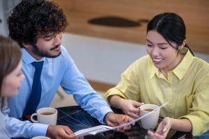 Two coworkers in a cafe-like setting review a document while sipping coffee at a table.