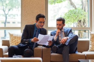 Two businessmen in suits sit on a beige sofa, reviewing documents together.