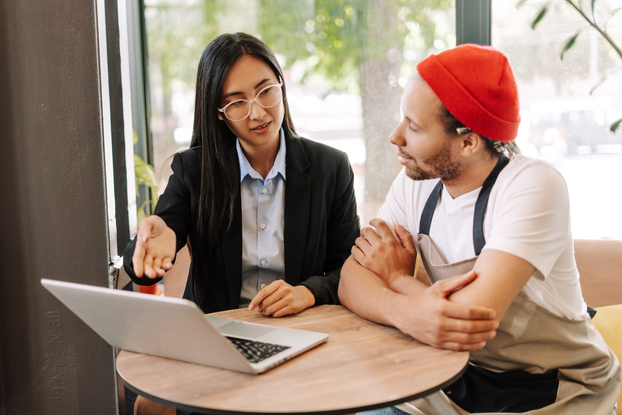 Two coworkers discuss something over a laptop at a wooden table in a bright cafe setting.